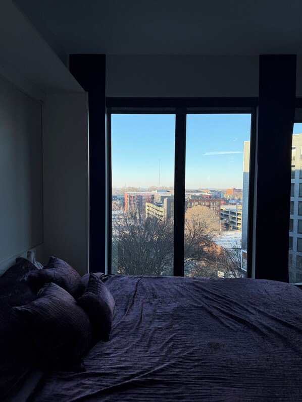 Un dormitorio con un ventanal grande, una cama con ropa de cama oscura y vistas a la ciudad.