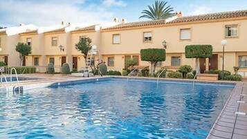Una piscina con solárium empedrado, rodeada por una hilera de edificios beige de arquitectura uniforme, con ventanas y puertas pequeñas.