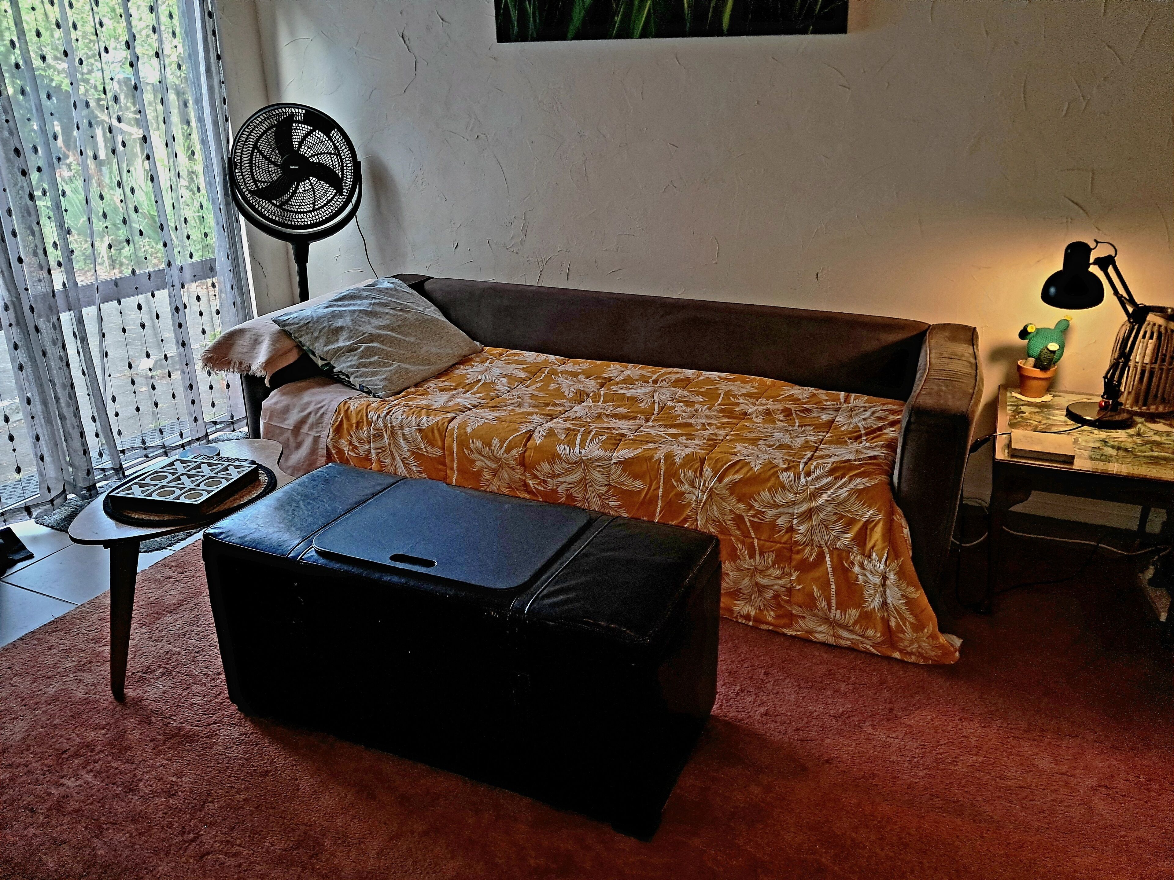 Un dormitorio pequeño con una cama individual, un escritorio, una silla, un ventilador de pie y una ventana con cortinas.