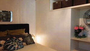 A bedroom with a bed, pillows, a bedside lamp, a wall-mounted shelf with storage boxes, and a decorative wall piece.