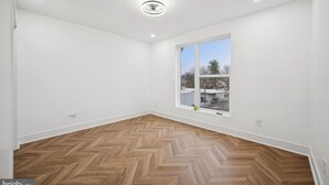 Ein geräumiges Zimmer mit großem Fenster, weißen Wänden und einem Fischgrätparkett.