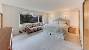 Ein Schlafzimmer mit einem großen Bett, einer Sitzbank mit Kissen, einem Nachttisch und einem Fenster mit Blick auf Bäume und ein Haus.