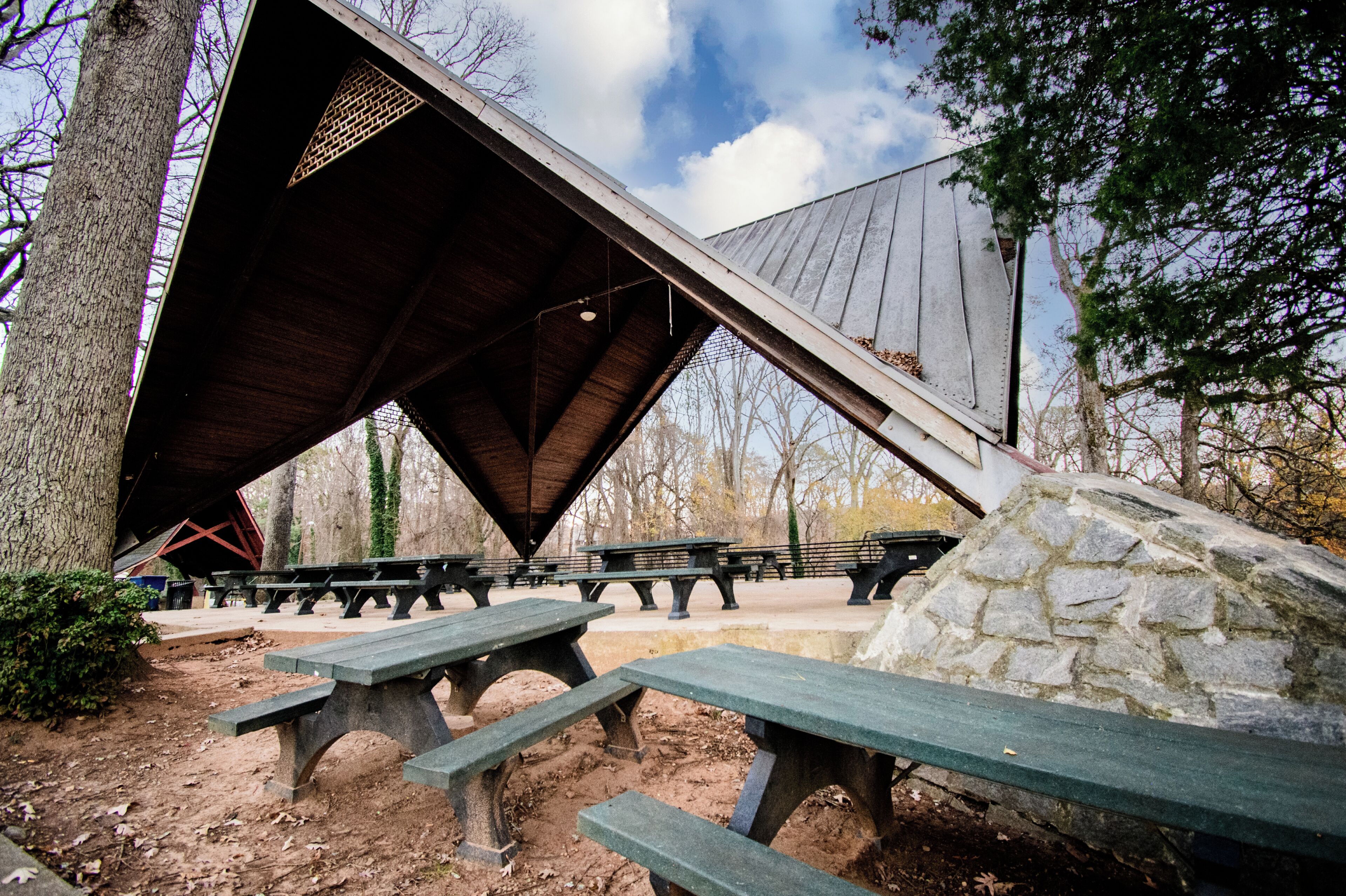 Un espace pique-nique abrité, doté de bancs et de tables à pique-nique, entouré d’arbres et d’un mur en pierre.