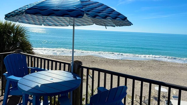 Un patio frente a la playa con una mesa, sillas y una sombrilla azul, con vista al océano.