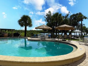 A swimming pool with a curved edge, surrounded by lounge chairs and umbrellas, with a view of trees and a body of water in the background. - Sunburst Gulffront – Beach, Bay & Gulf Views (Englewood)