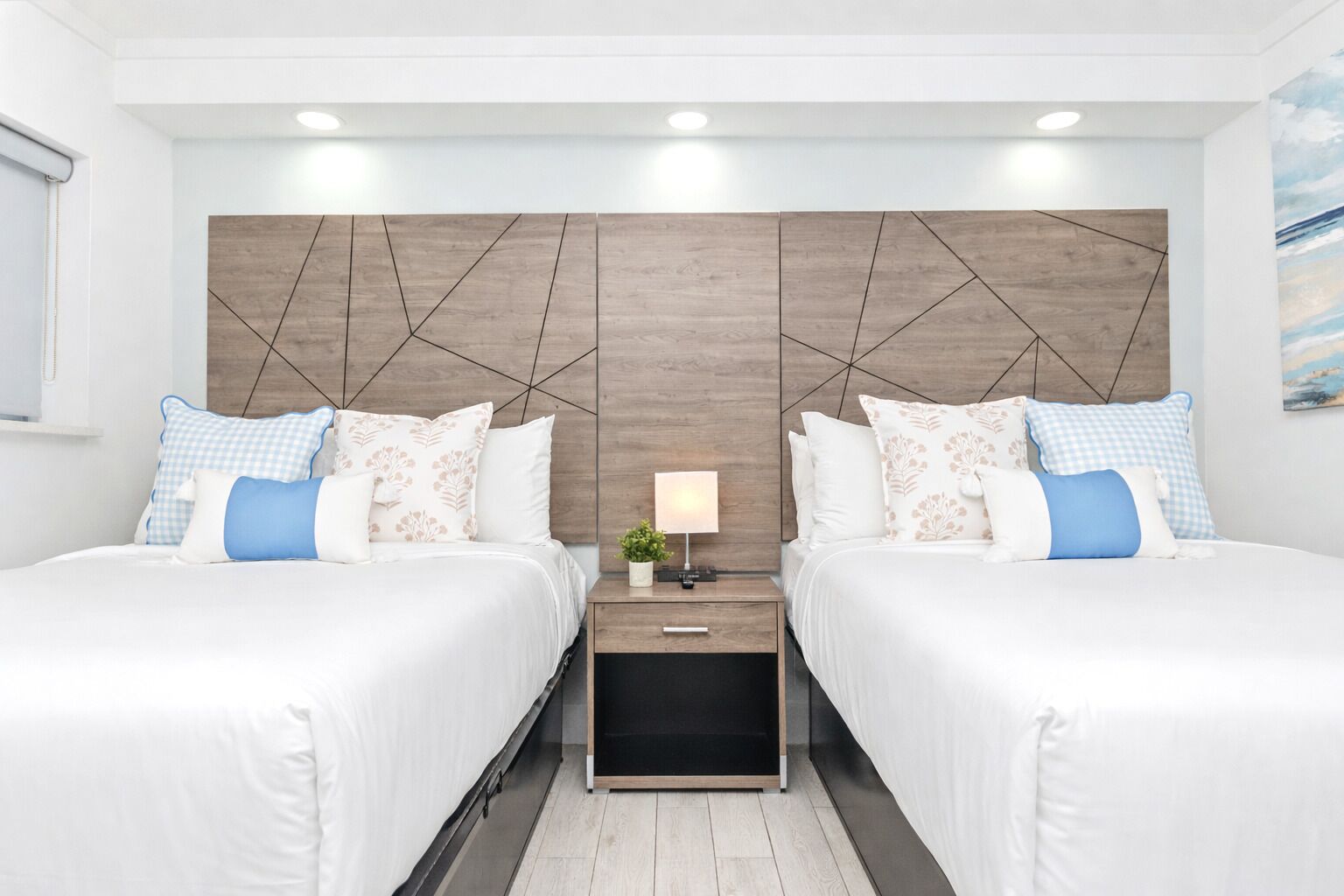 Quarto de hotel com duas camas, uma mesa de cabeceira com abajur e cabeceira de madeira.
