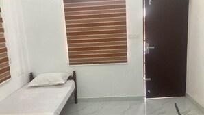Une petite pièce propre, avec un lit simple, une fenêtre avec des stores, une porte et un interrupteur sur le mur.