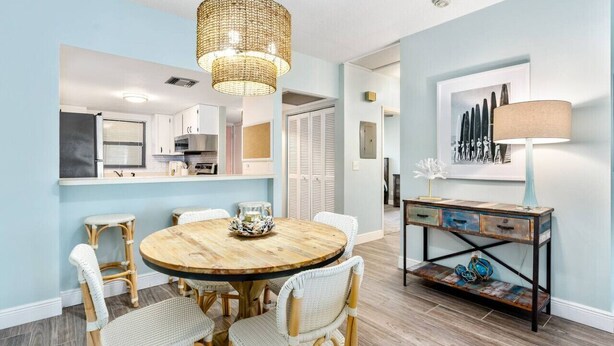 A dining area with a round wooden table surrounded by wicker chairs, a woven pendant light, and a rustic console table with a lamp. - Island Time Oasis Pool Walk to Beach & Balcony (Daytona Beach)