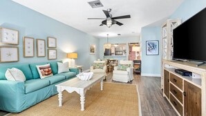 A living room with a teal sofa, white coffee table, and a TV on a wooden cabinet. - Island Time Oasis Pool Walk to Beach & Balcony (Daytona Beach)