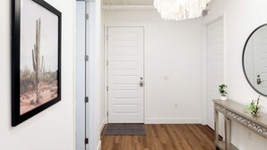 Un pasillo con una puerta blanca, un cuadro en la pared, suelo de madera, una mesa consola con un espejo y una lámpara colgante.