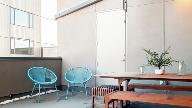 Un espacio exterior moderno con un banco de madera y dos sillas azules, una mesa pequeña con una planta, y un suelo de hormigón.