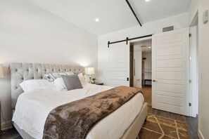 Un dormitorio con una cama grande, un cabecero, una mesita de noche con lámpara y una puerta corredera a otra habitación.