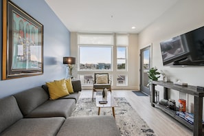 A modern living room with a grey sofa, a coffee table, a TV, and a large window offering a city view. - Golden Hour Designer Loft Espadin Lohi (Denver)