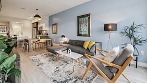 A modern living room with a grey sofa, wooden armchairs, a coffee table, and a dining area with a kitchen in the background. - Golden Hour Designer Loft Espadin Lohi (Denver)