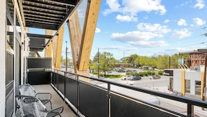 Een balkon met metalen stoelen, uitzicht op een straat met auto's en gebouwen, en een heldere lucht.