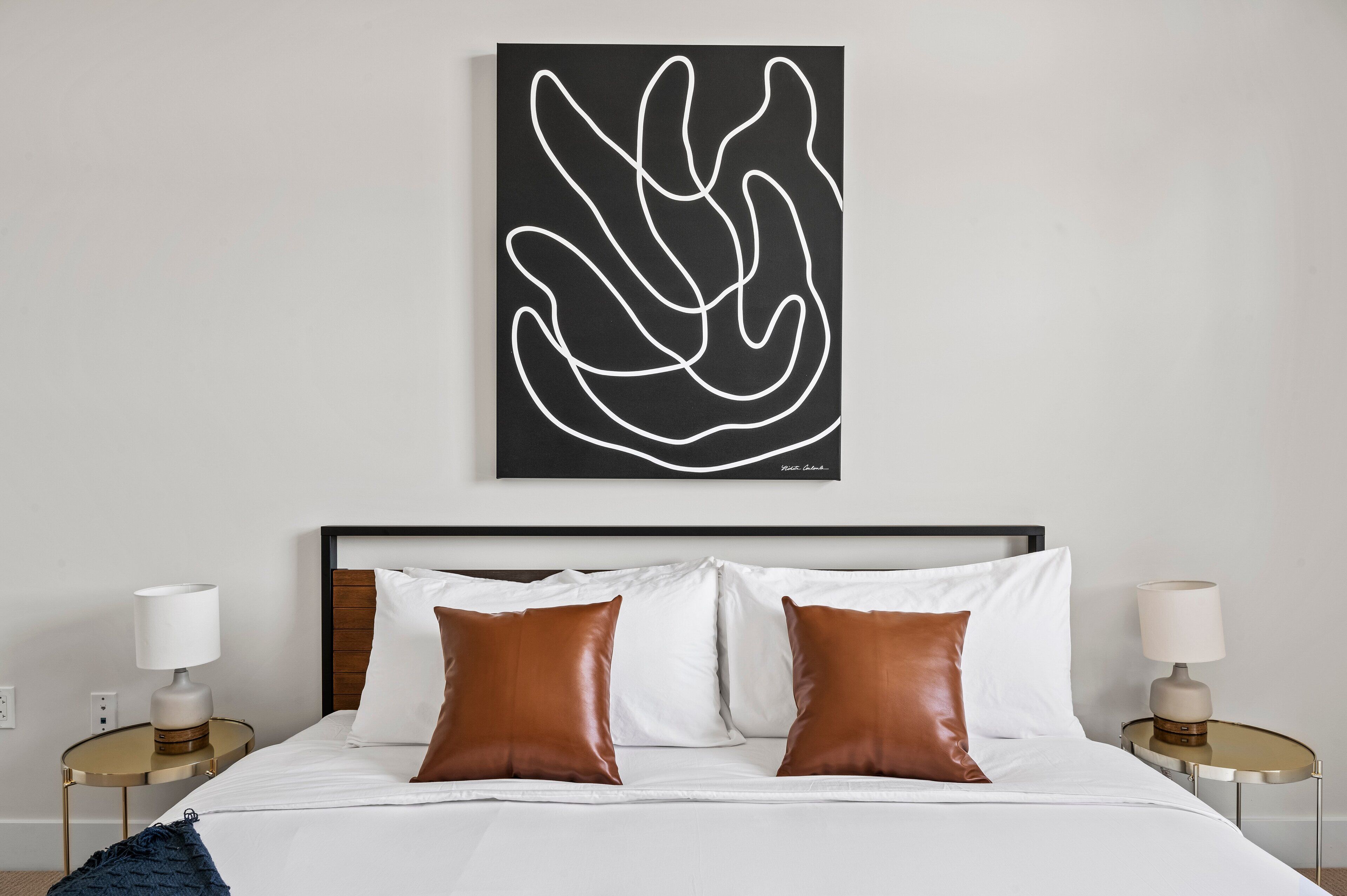 A modern hotel room with a large bed, two bedside tables with lamps, and a black abstract artwork on the wall.