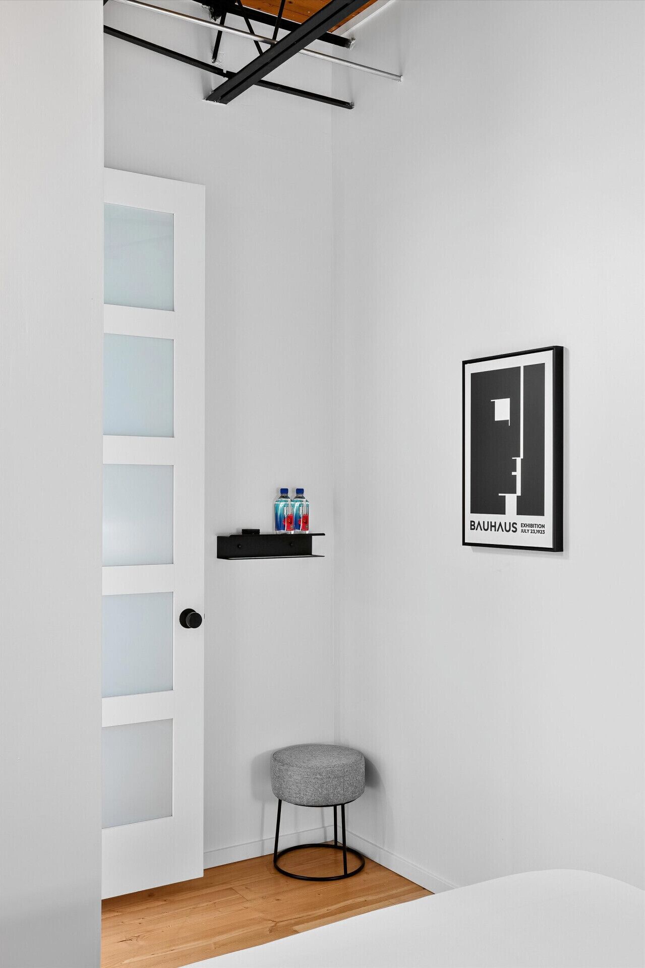 A minimalist room with a white door, a black metal shelf with water bottles, a grey ottoman, and a framed Bauhaus exhibition poster.