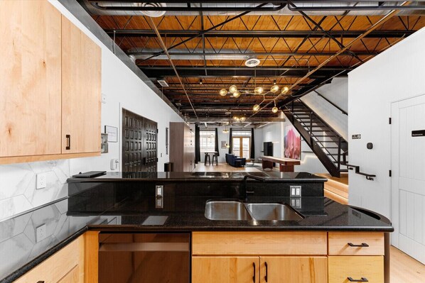A modern kitchen with a black countertop, wooden cabinets, and a visible staircase leading to another level. - Suite [1] + [2] (Denver)
