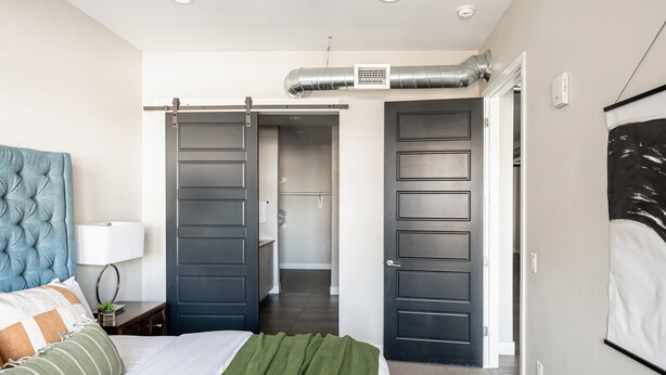Ein Schlafzimmer mit Bett, Nachttisch, Lampe, großem Fenster und einer Schiebetür zu einem Badezimmer.