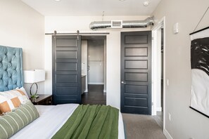 Ein Schlafzimmer mit Bett, Nachttisch, Lampe, großem Fenster und einer Schiebetür zu einem Badezimmer.
