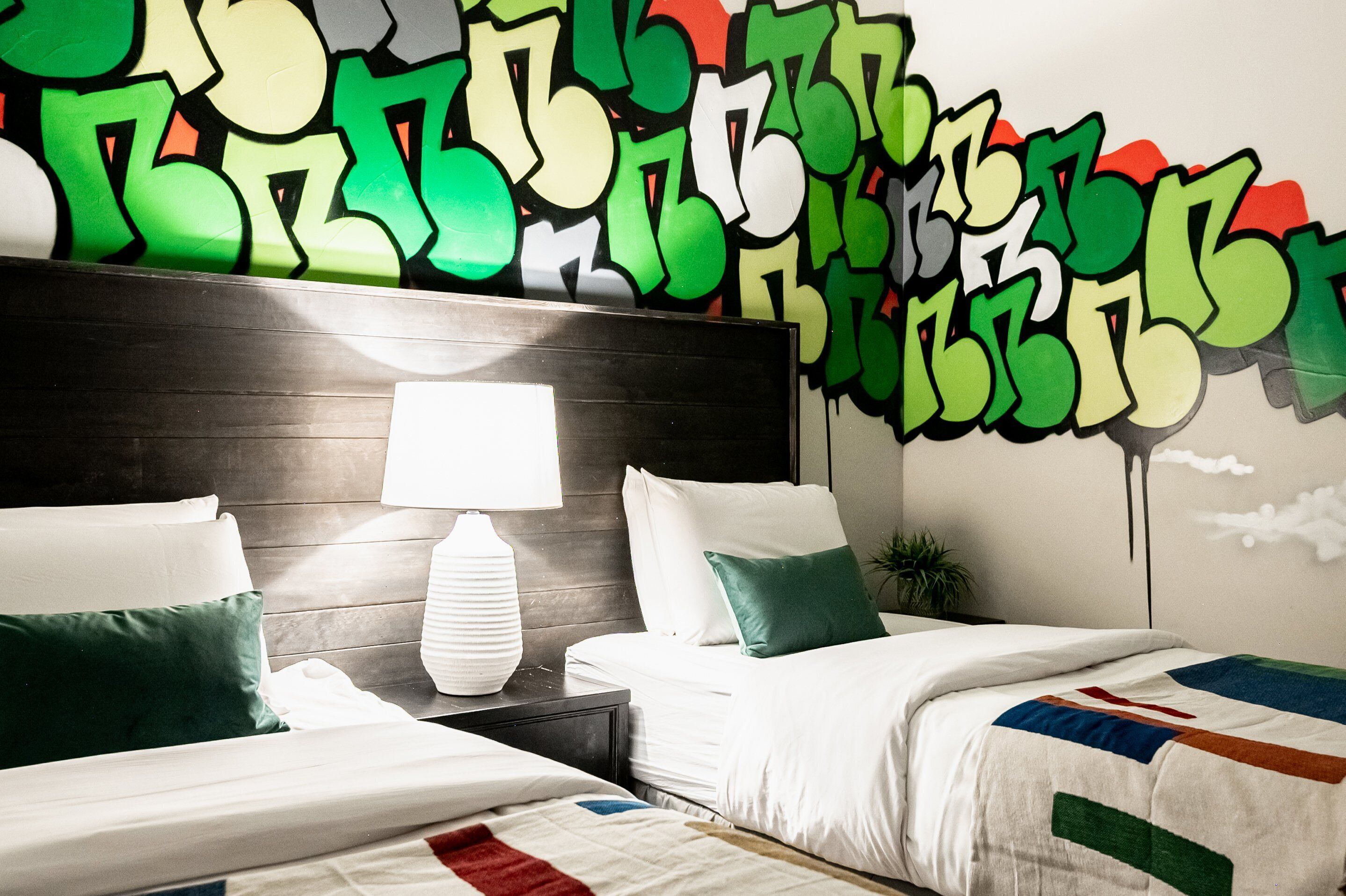 A hotel room with a bed, a nightstand, a lamp, and a wall with graffiti art.
