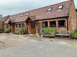 A brick building with a tiled roof, a wooden door, and multiple windows. There are potted plants and a bench outside.