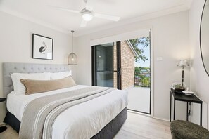 Un dormitorio moderno con cama, ventilador de techo, una obra de arte enmarcada, puerta corrediza de cristal y una mesita de noche con lámpara.