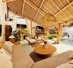 A spacious outdoor lounge area with a thatched roof, comfortable seating, and a pool. - Casa Alonia (Uluwatu)