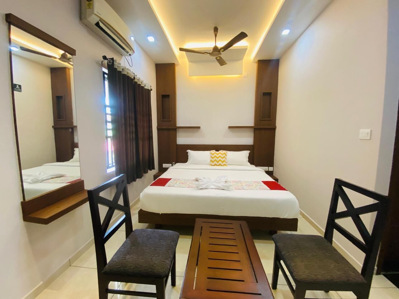 A hotel room with a bed, two chairs, a bench, a ceiling fan, and an air conditioning unit.