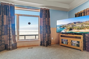 A room with a large window, a flat-screen TV, and a wooden entertainment unit with books. - Sandy Bottom: Summer Beachfront Bliss | Swim, Kayak, Relax (Ocqueoc)