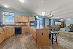 A modern kitchen with wooden cabinets, a central island, and a dining area with a table and chairs. - Sandy Bottom: Summer Beachfront Bliss | Swim, Kayak, Relax (Ocqueoc)