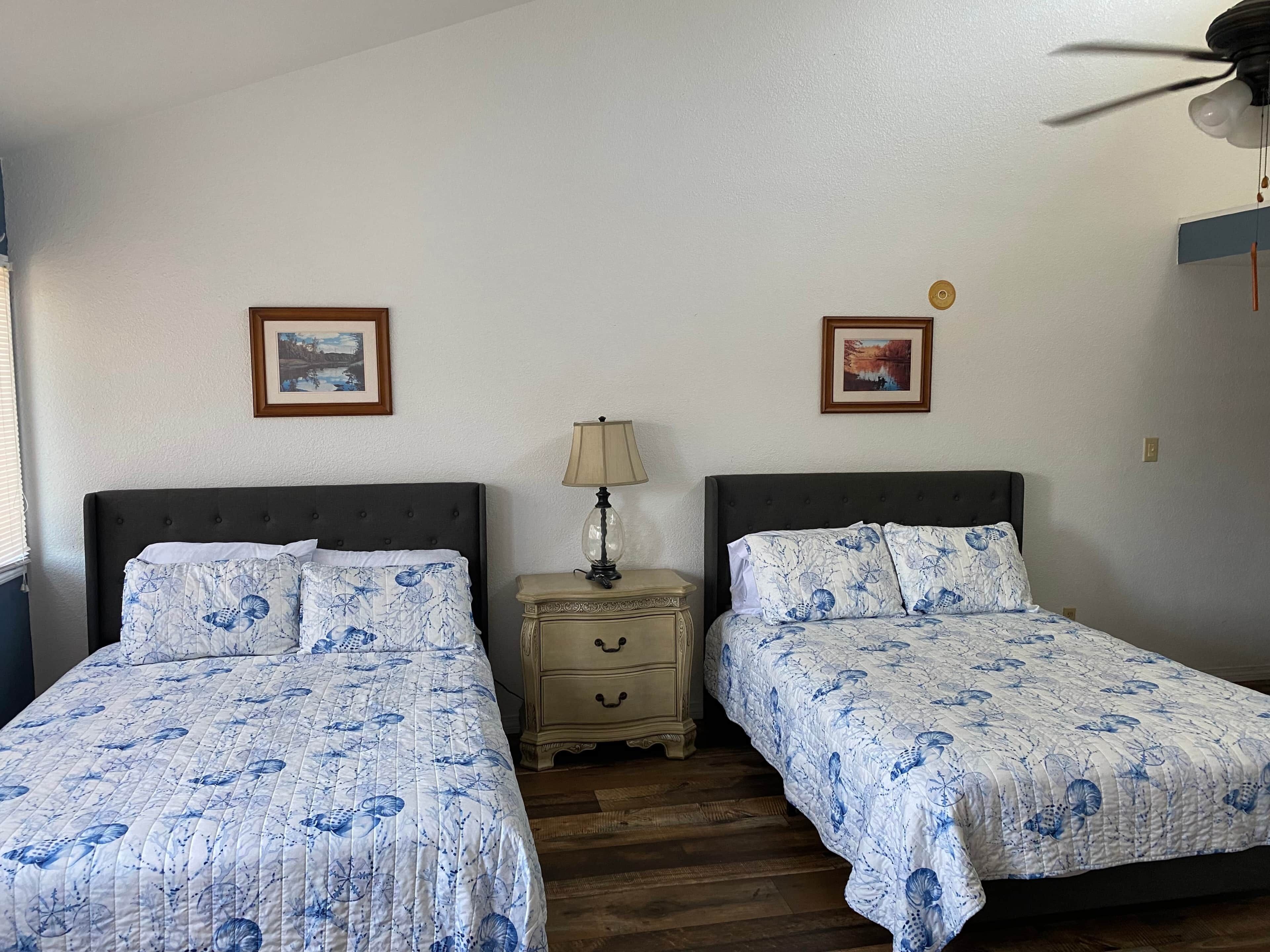 A room with two beds, each with a floral patterned bedspread, a nightstand with a lamp, and two framed pictures on the wall.