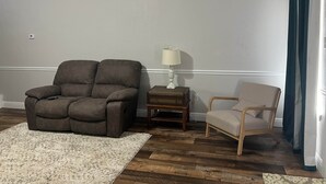 Ett vardagsrum med en brun soffa, ett litet träbord med en lampa, en beige fåtölj och en mönstrad matta.