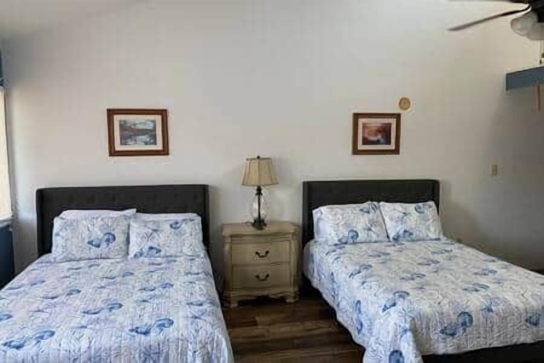 A room with two beds, each with a patterned bedspread, a bedside table with a lamp, and framed pictures on the wall.
