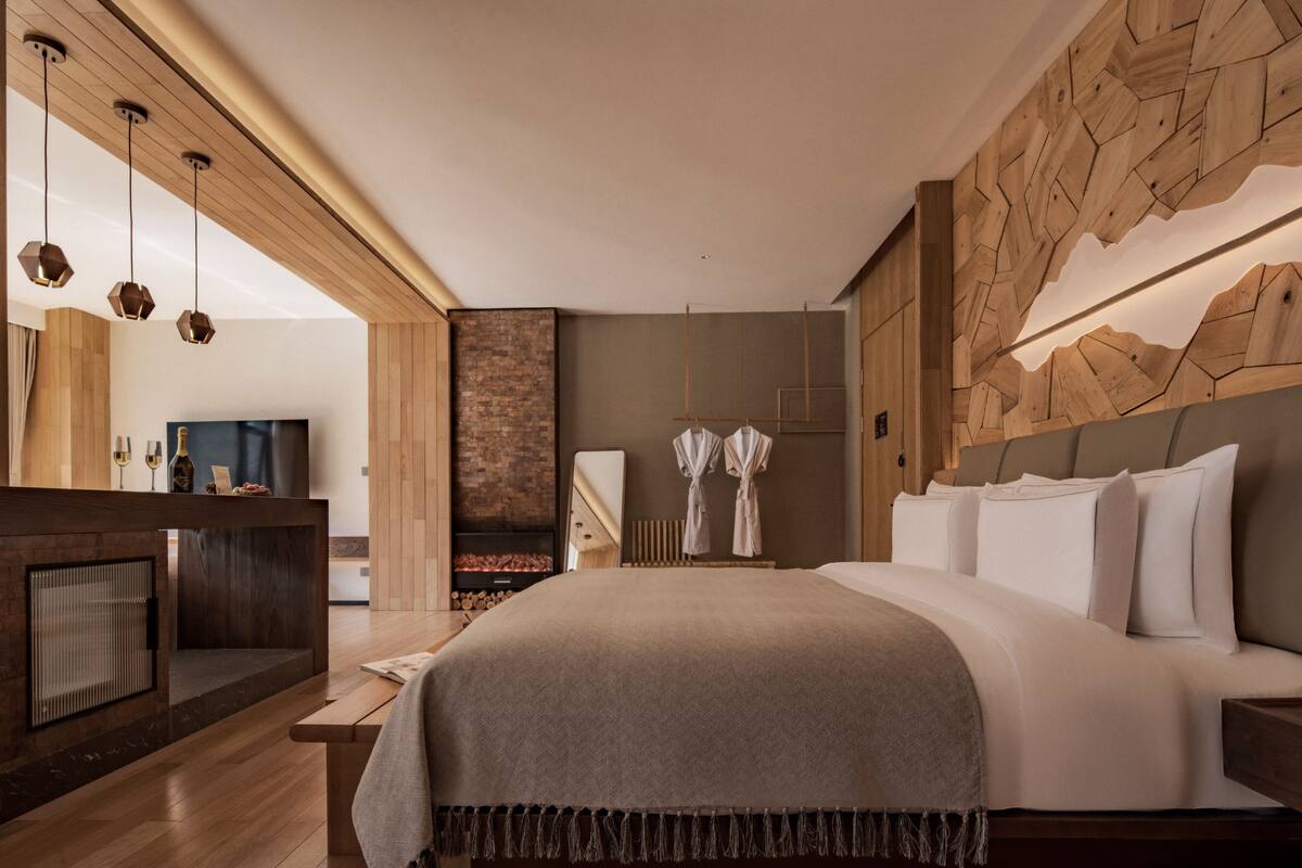 A modern hotel room with a large bed, a wooden headboard, a fireplace, a TV, and a stone wall.