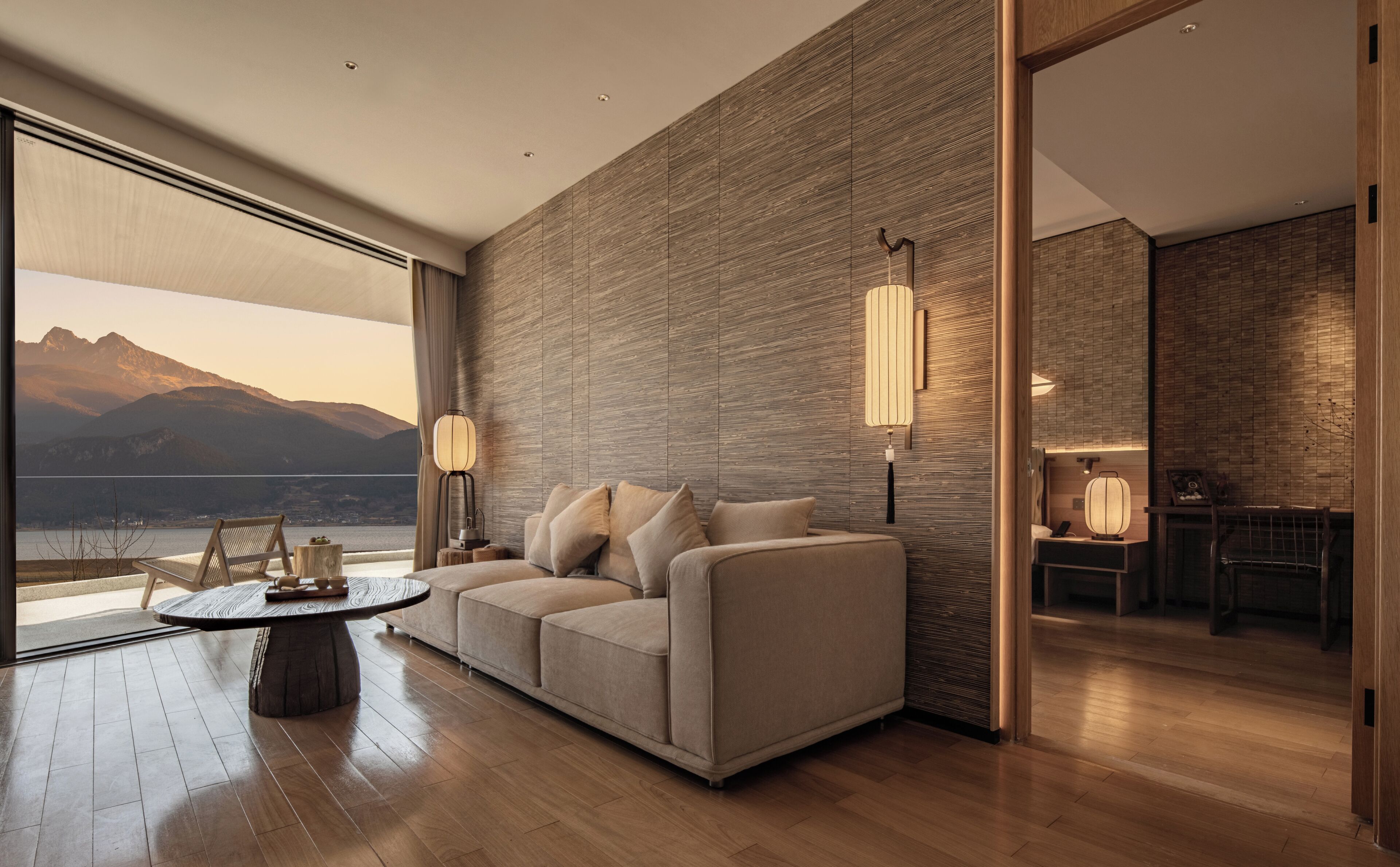 Una sala de estar moderna con un sofá, una mesa de centro y dos lámparas de pared. Un ventanal ofrece vistas a montañas y a un lago.