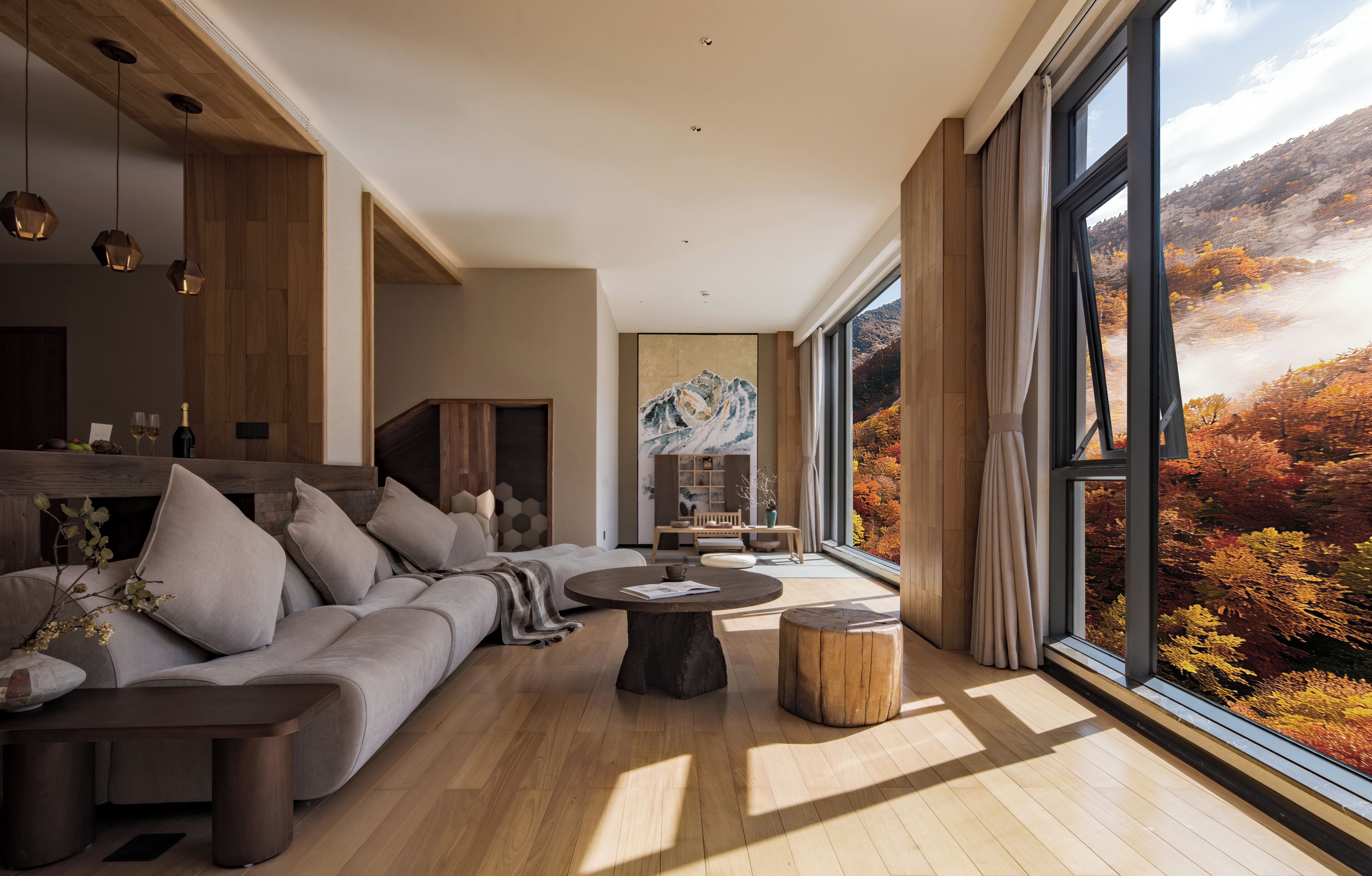 A modern living room with a large sectional sofa, a round coffee table, and a view of autumn foliage through large windows.