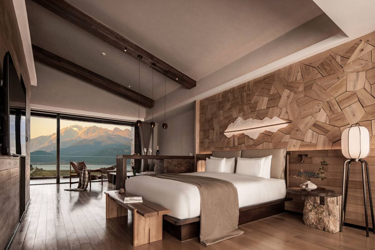 A modern bedroom with a large bed, wooden headboard, a view of mountains, and a wooden floor.