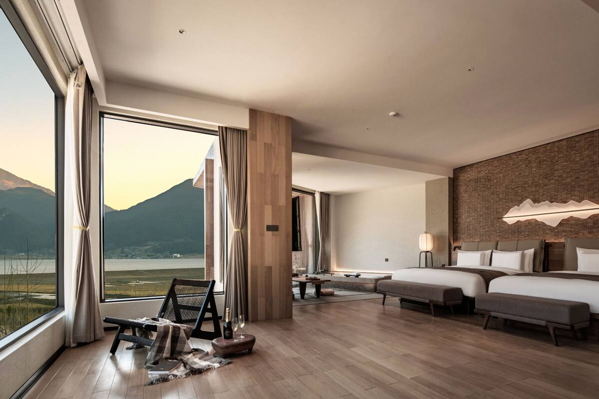 A modern hotel room with a large window, a bed, a sofa, a small table, and a lamp.