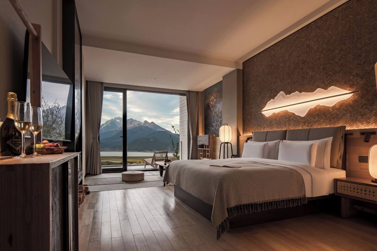 A modern hotel room with a large bed, a desk with a bottle and glasses, a view of mountains, and a unique wall light.