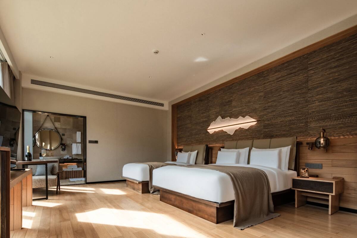 A modern hotel room with a large bed, a wooden headboard, a bedside table, a mirror, and a wooden floor.
