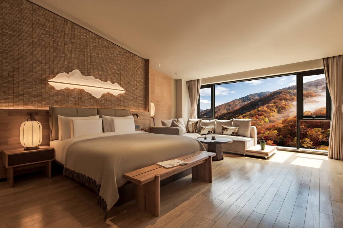 A modern hotel room with a large bed, a sofa, a small table, a nightstand, and a large window with a mountain view.