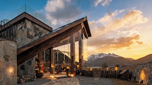 Un chalet de montagne aux murs en pierre, doté d’un patio vitré et offrant une vue sur des montagnes enneigées.