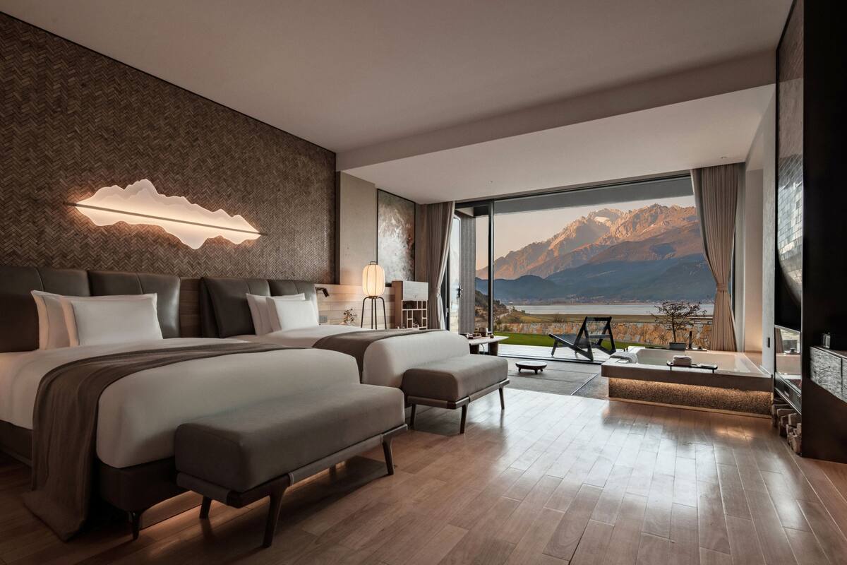 A modern hotel room with a large bed, a second smaller bed, a seating area, and a view of mountains through large windows.