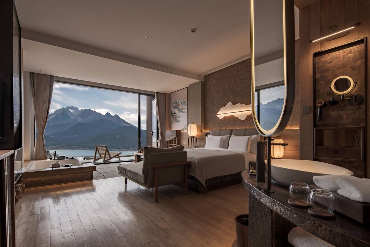 A modern hotel room with a large window, a bed, a sitting area, and a bathroom.
