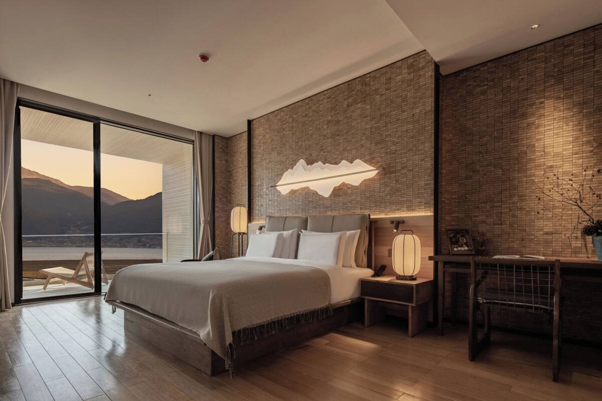 A modern bedroom with a large bed, a nightstand, a desk, and a view of the outdoors.