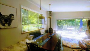 A dining area with a long table, chairs, and a sofa. There are large windows with a view of greenery outside. - Caravan and house set up for mountain biking (Warburton)