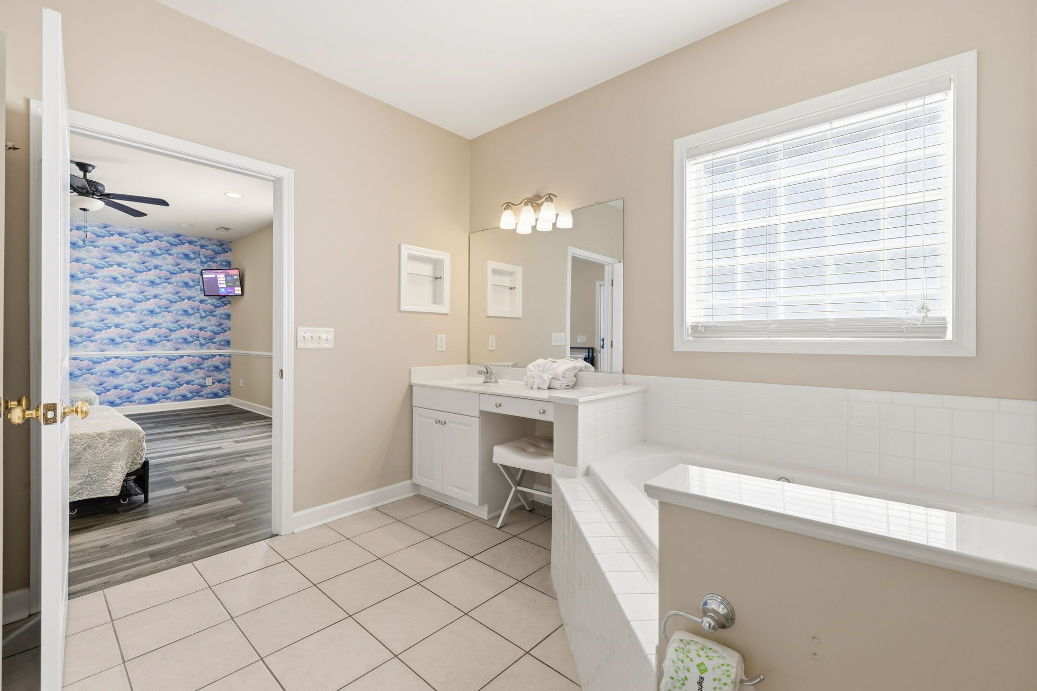 A bathroom with a vanity, a mirror, a window with blinds, a ceiling fan, and a wall-mounted TV.
