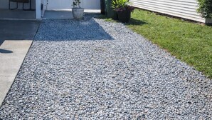Een woonhuis met een grindpad, een witte schutting en een kleine tuin met potplanten.