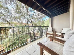 Un balcón con una silla y una mesa de madera, baranda metálica y vistas a árboles y edificios.